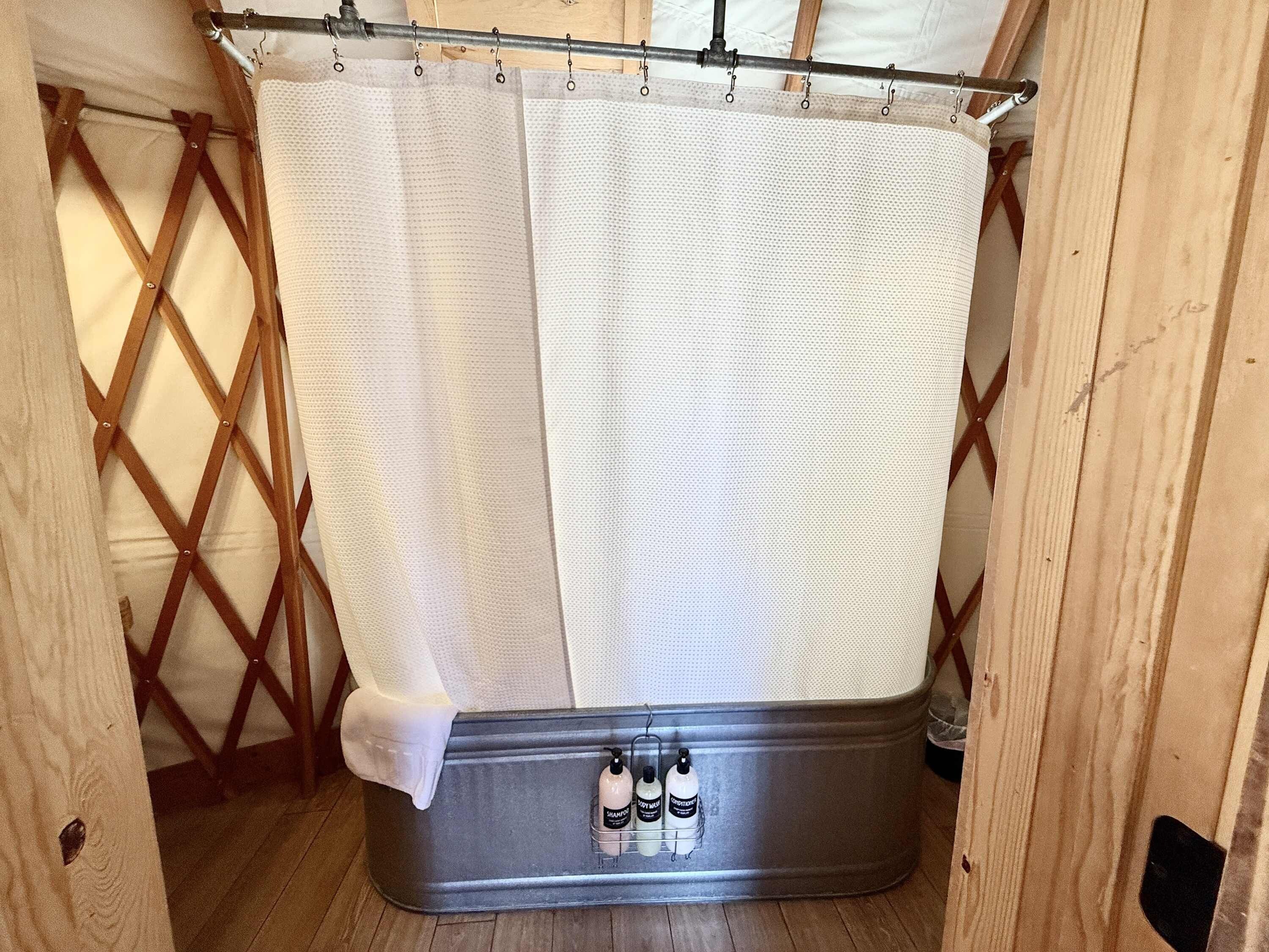 A shower area with a metal tub, a wooden lattice wall, and a shower curtain. There are three bottles of shampoo and conditioner on the tub.
