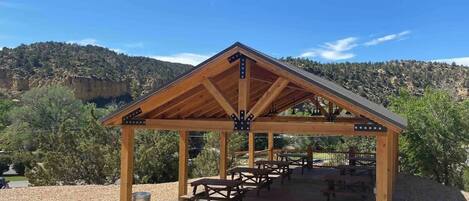 A wooden pavilion with picnic tables and trash bins, set against a mountainous backdrop.