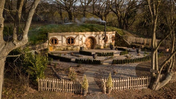 A rustic stone building with circular windows and a chimney emitting smoke, surrounded by a wooden fence and a gravel pathway. - Oastbrook Estate Vineyard (Robertsbridge)