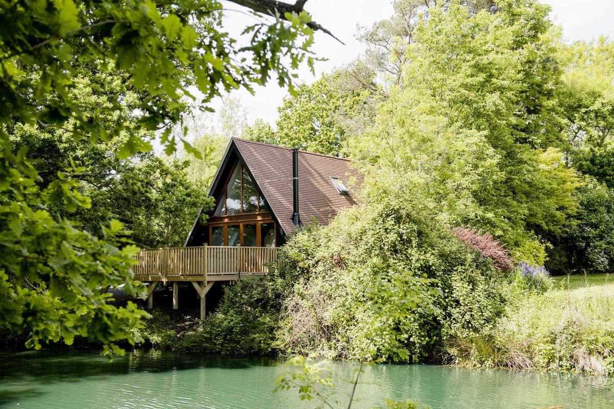 A cabin with a wooden deck and large windows overlooking a lake, surrounded by dense greenery.