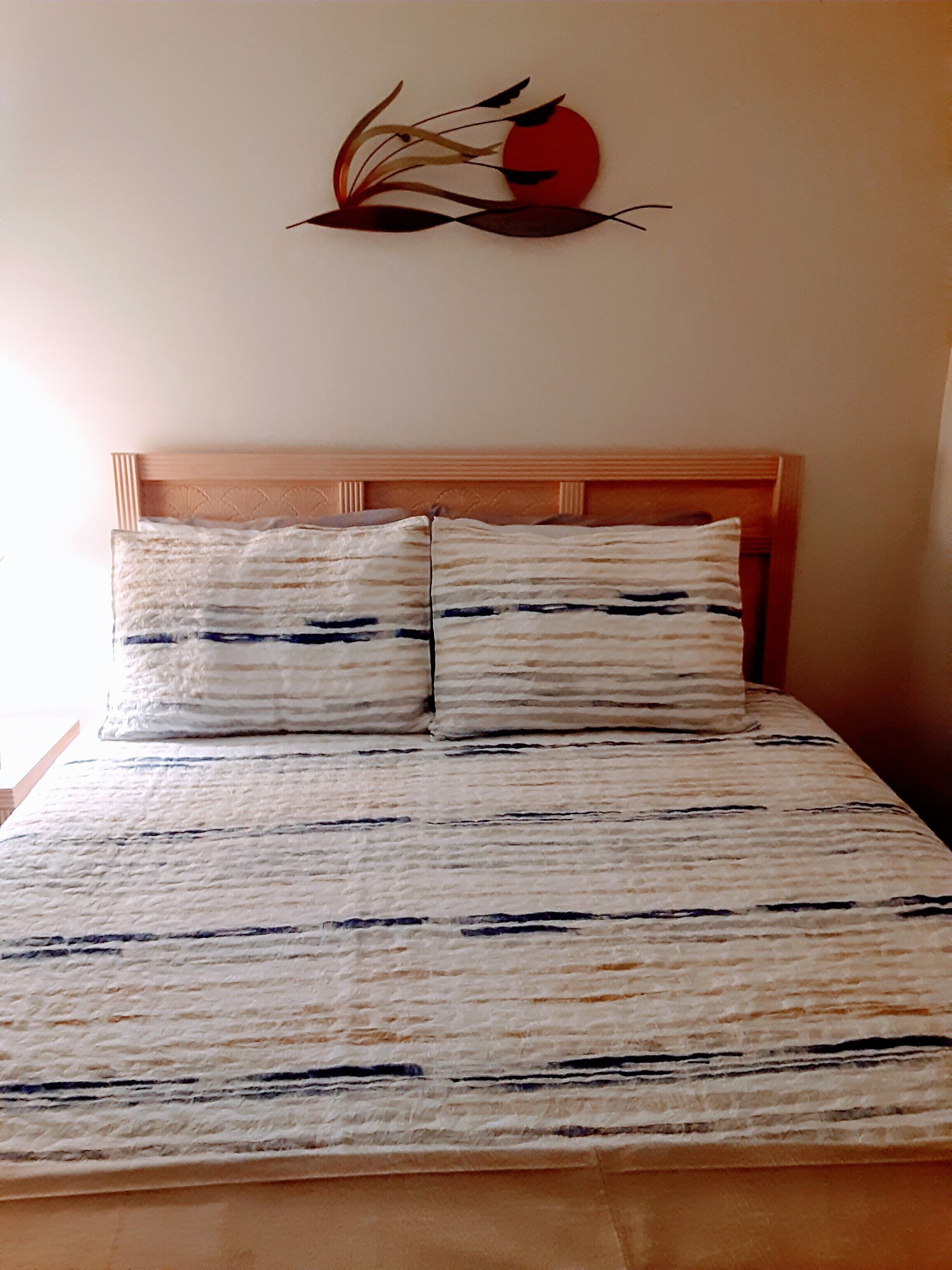 A bed with a striped bedspread and two pillows, a wooden headboard, and a wall art piece above the bed.