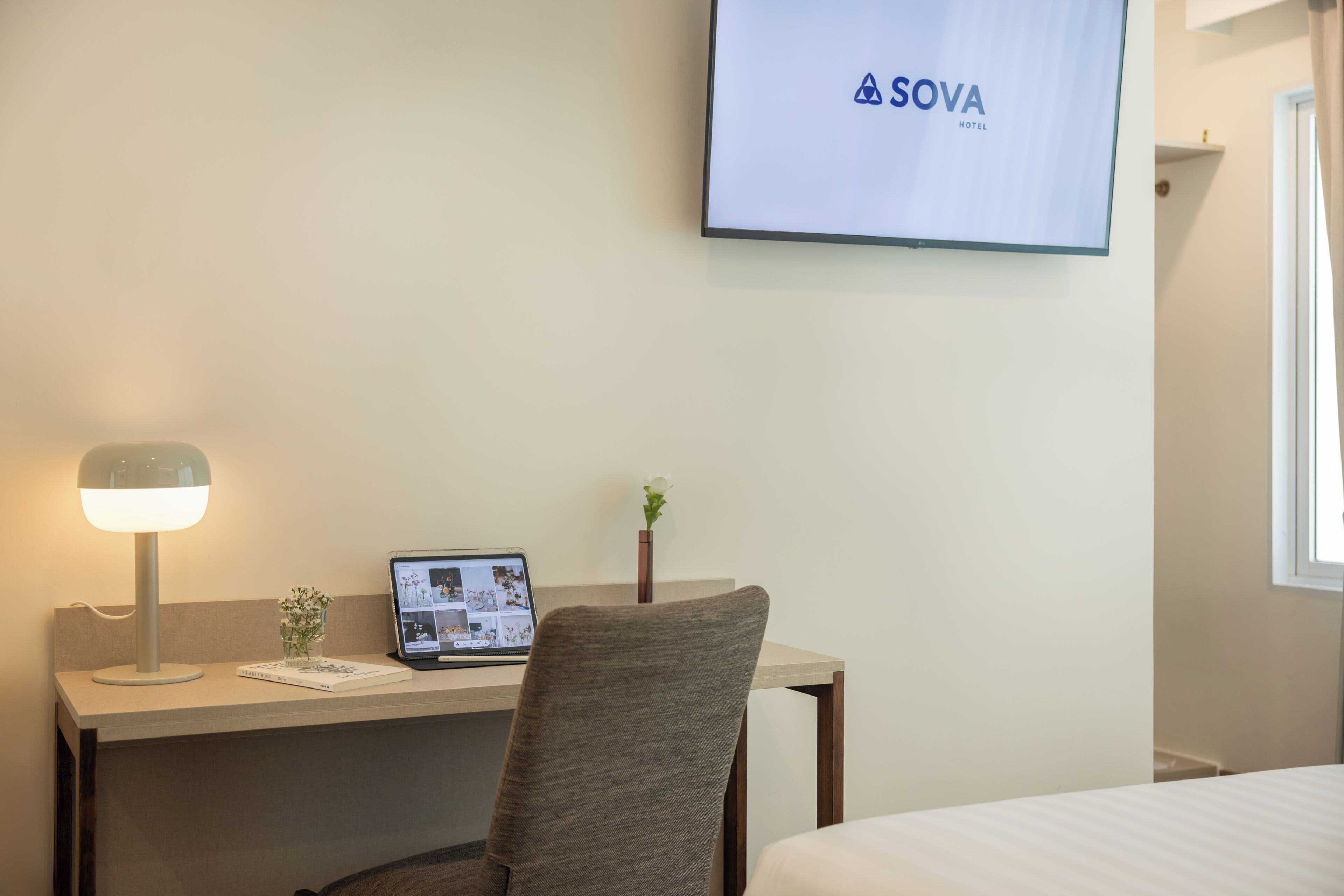Een hotelkamer met een bureau, een laptop, een stoel, een bed, een televisie waarop het logo van het hotel staat, en een raam.