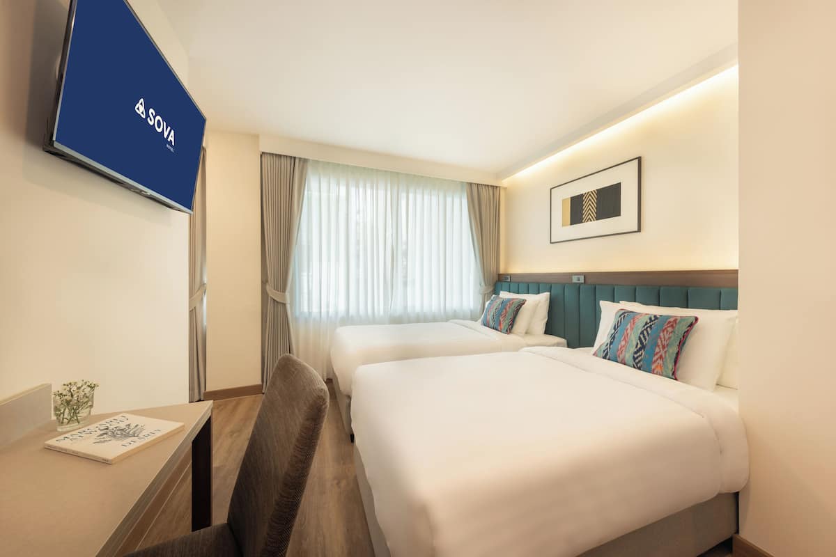 A hotel room with two beds, a TV mounted on the wall, a desk with a chair, and a window with curtains.