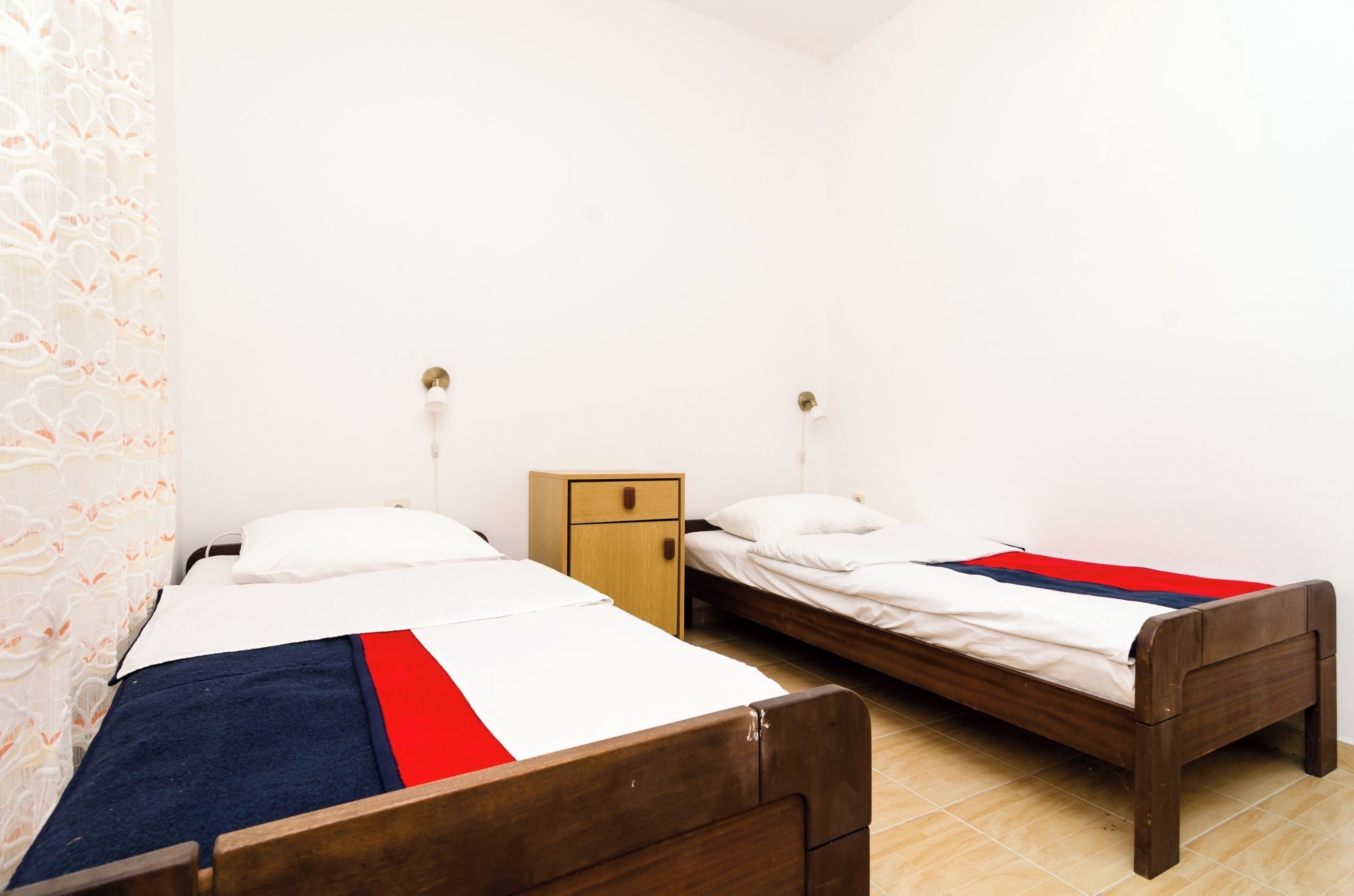 Quarto com duas camas de solteiro, uma mesa de cabeceira de madeira e uma luminária fixada na parede.