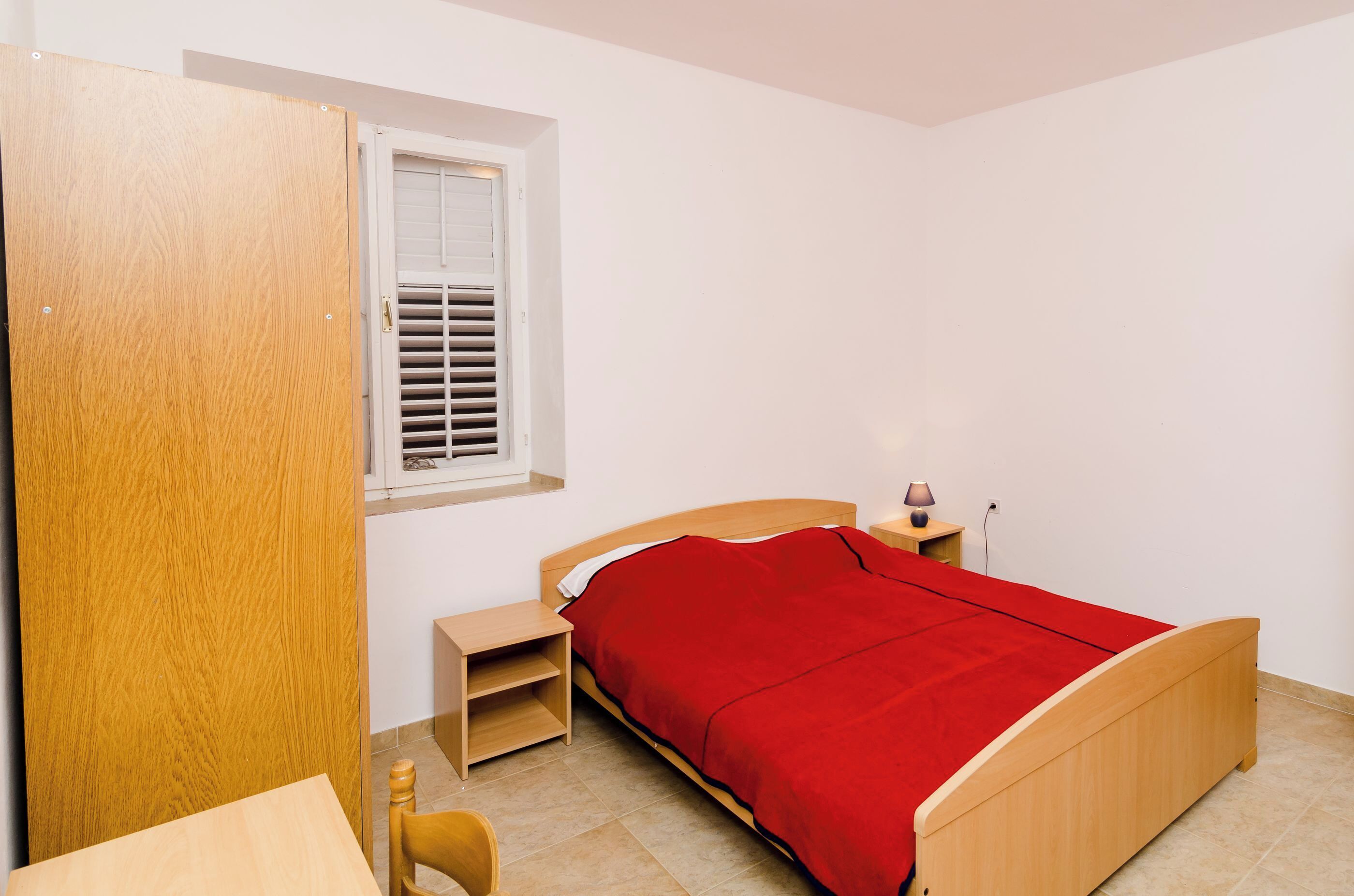 Un dormitorio con cama de madera, colcha roja, mesita de noche con lámpara y ventana con persianas.