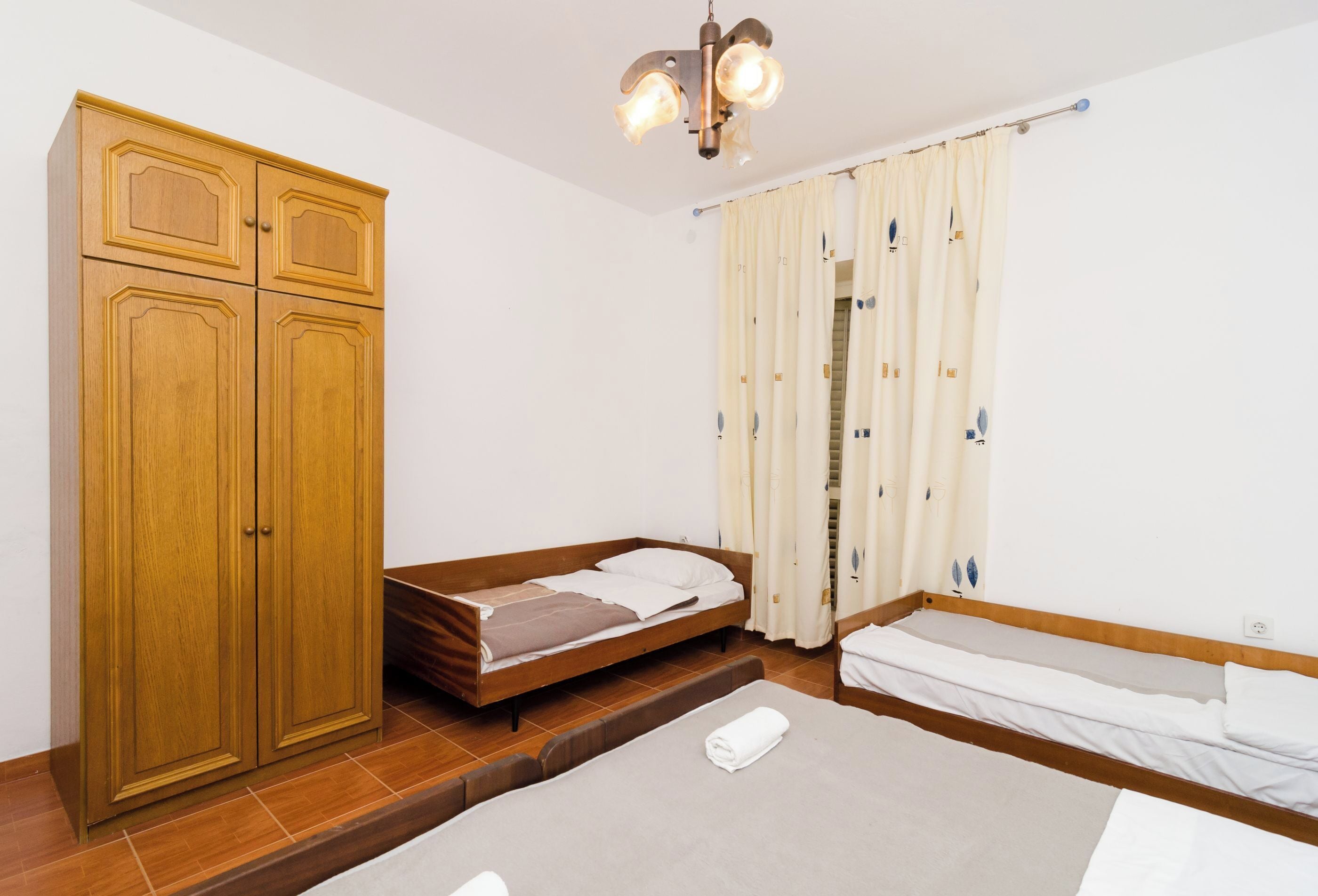 Un dormitorio con un armario de madera, una cama con colchón y almohadas, una mesita de noche y una lámpara en el techo.