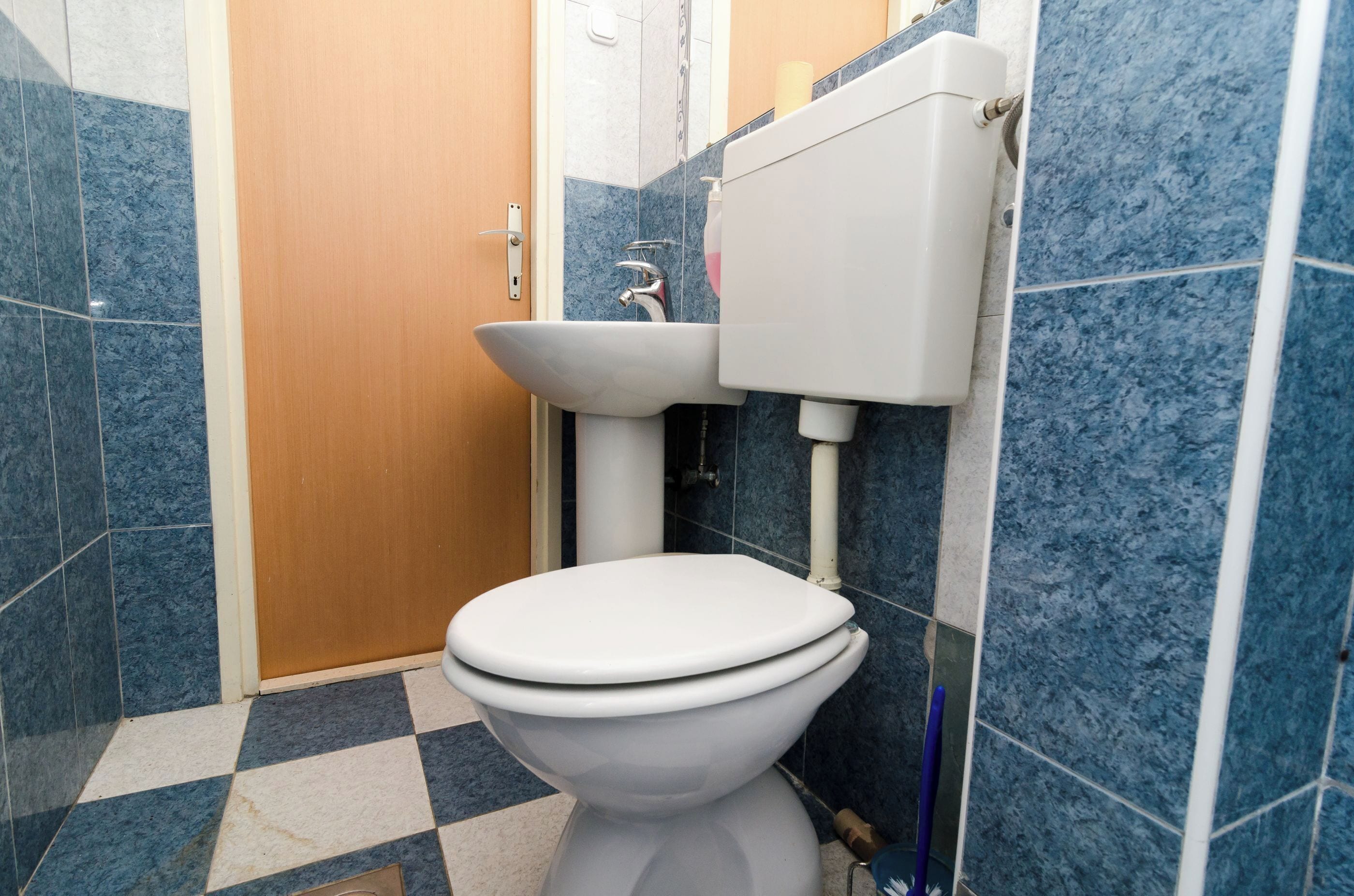 Et badeværelse med et hvidt toilet, en vask og en vandbeholder. Væggene er flisebelagt i blå og hvid, og der er en trædør.