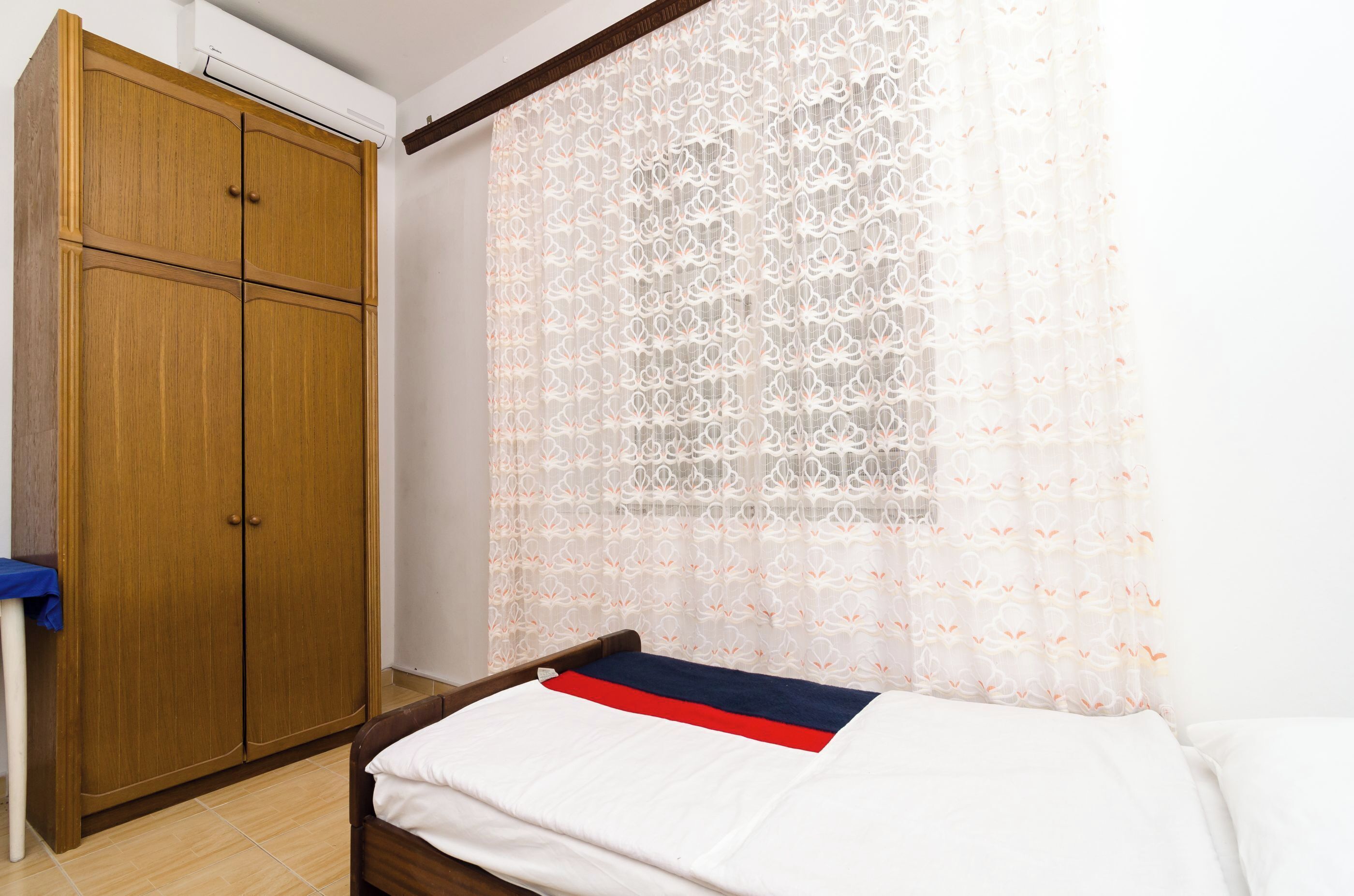 A bedroom with a wooden wardrobe, a bed with a white and red blanket, and a sheer curtain with a floral pattern.