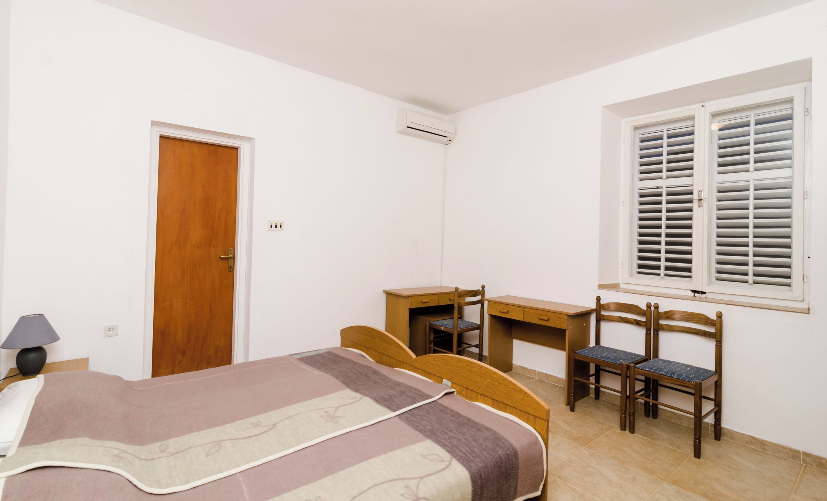 Ein Schlafzimmer mit einem Bett, einem Schreibtisch, zwei Stühlen, einem Fenster mit Rollläden, einer Tür und einer Klimaanlage.
