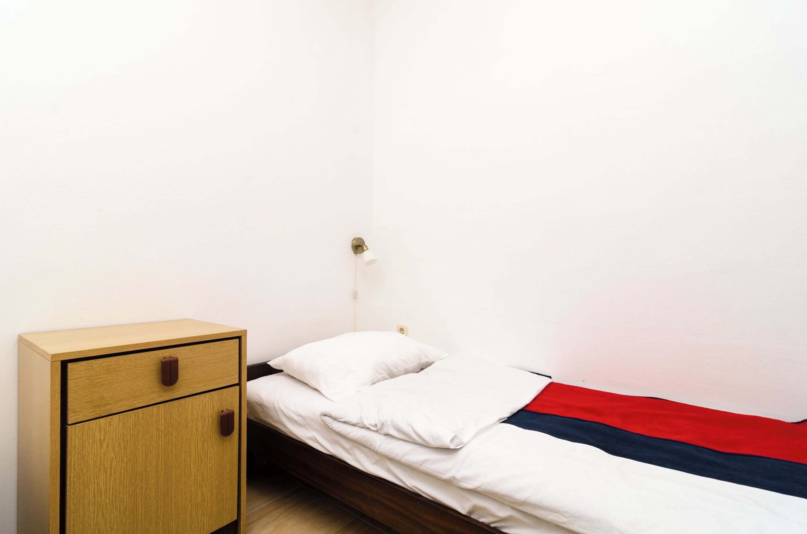A small, single-bed room with a wooden nightstand and a red, white, and blue blanket on the bed.