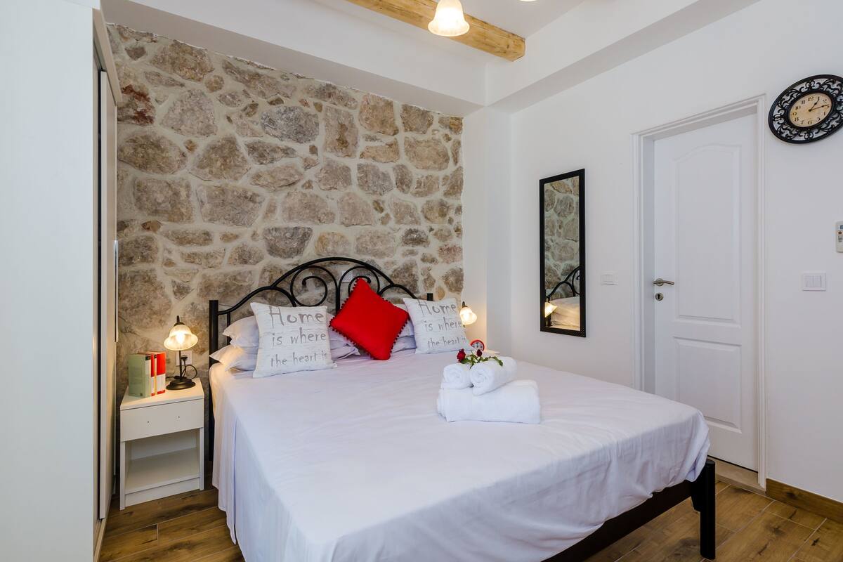A bedroom with a stone wall, a bed with white bedding, a red pillow, and a clock on the wall.