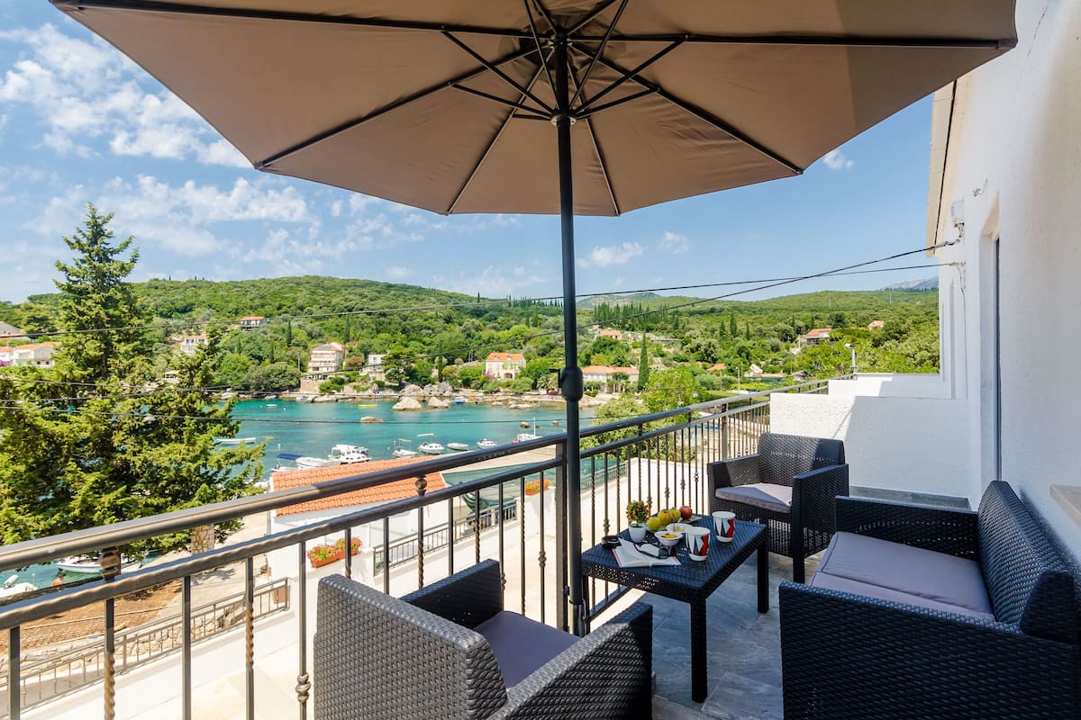 A balcony with wicker furniture, a table with a vase, and a view of a coastal town and water.