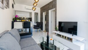 Een moderne woonkamer met een grijze bank, een glazen salontafel, een flatscreen televisie en een keuken op de achtergrond.