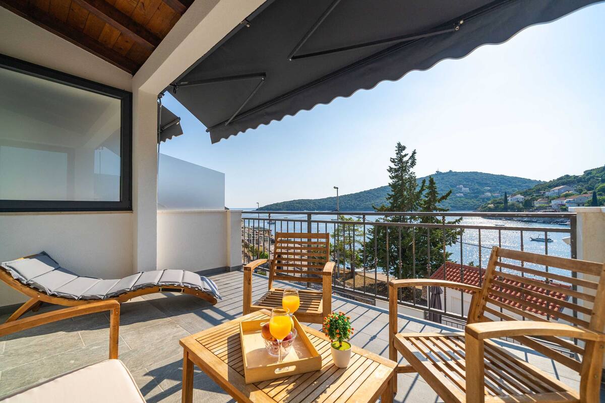 A balcony with wooden furniture, a tray with drinks, and a view of a body of water and hills.