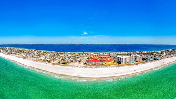 An aerial view of a coastal resort with a long beachfront, clear blue waters, and a variety of buildings including a prominent red-roofed hotel.