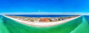 Ett flygbildsbild av en kustort med en lång strand, kristallklart blått vatten och en mängd olika byggnader, inklusive ett framträdande hotell med rött tak.