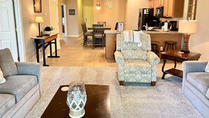 A living room with a sofa, a coffee table, and a patterned armchair. There is a kitchen area with wooden cabinets and a refrigerator.