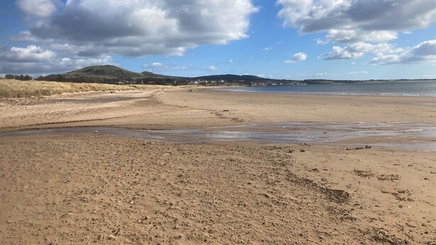 Una ampla platja sorrenca amb ones suaus, cel blau i turons allunyats.