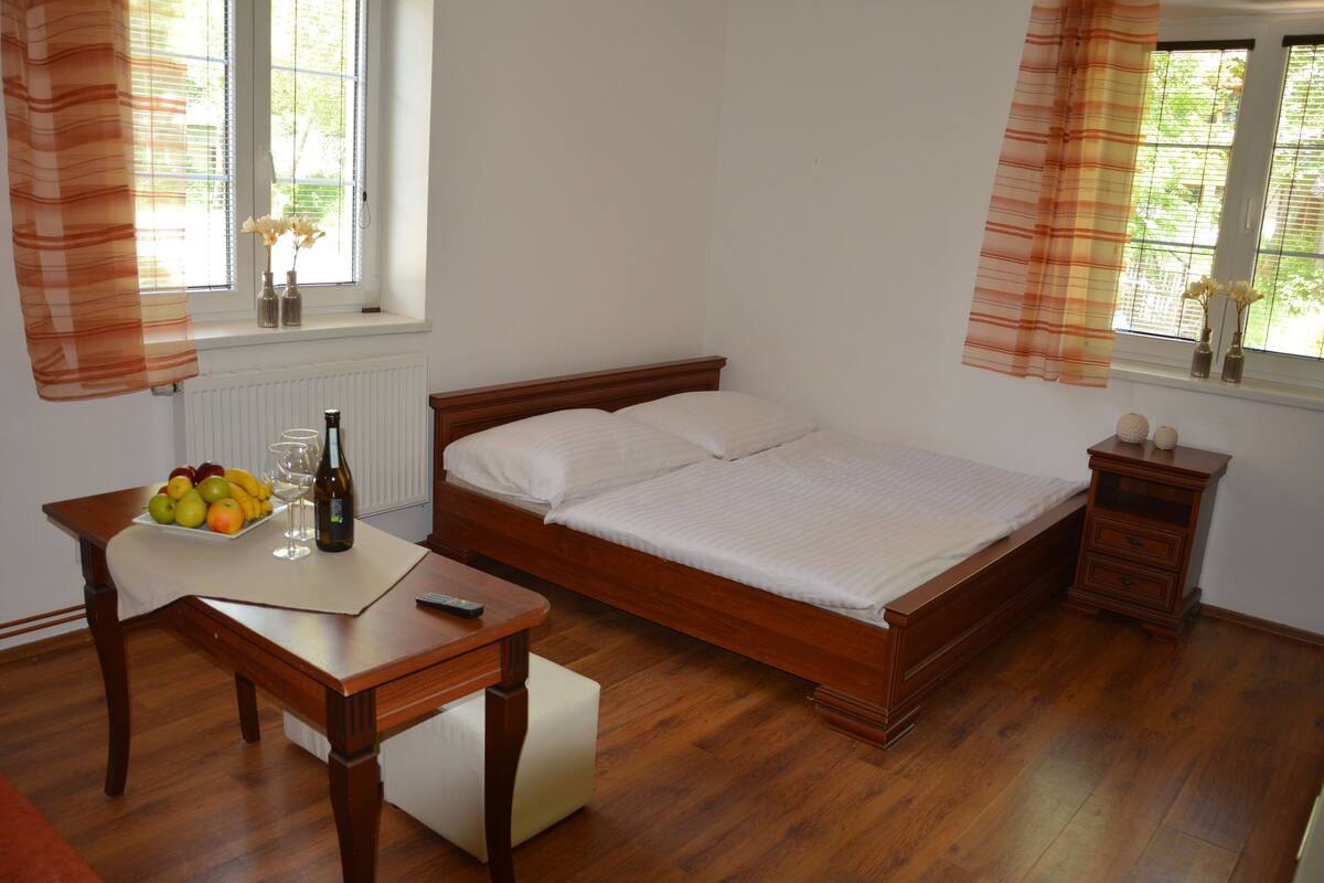 Une chambre à coucher avec un lit en bois, une table de chevet sur laquelle se trouvent un panier de fruits, une bouteille et une télécommande. Il y a une fenêtre avec des rideaux et un radiateur en dessous.