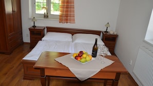 A neatly made bed with white linens, a wooden nightstand with a lamp, a bedside table with a fruit bowl and wine bottle, and a window with curtains. - Apartmány Divoký anděl (Dolní Moravice)