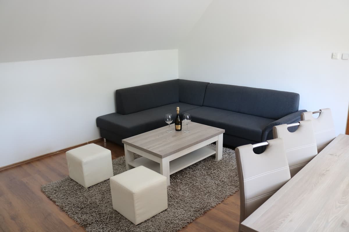 Un salon moderne avec un canapé d’angle gris foncé, une table basse en bois clair et deux poufs blancs.