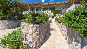 A building with a stone pathway leading to a covered entrance, surrounded by lush greenery and flowering plants. - Apartments Djurkovic (Zupa dubrovacka)