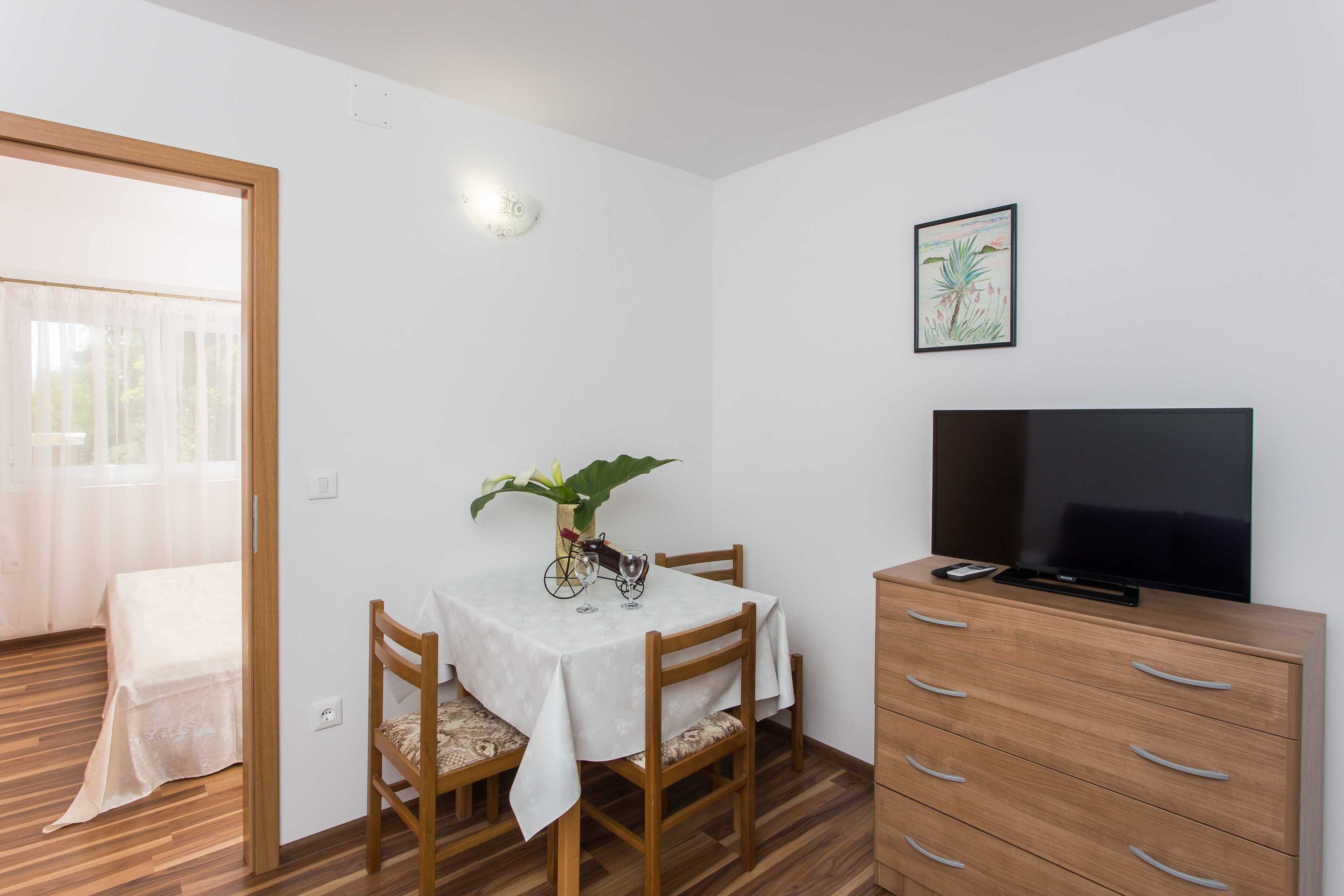 A small, well-lit room with a wooden floor, a dining table with chairs, a TV on a wooden cabinet, and a window with sheer curtains.
