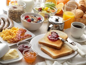 Una mesa de desayuno con diversos alimentos, como tostadas con mermelada, huevos revueltos, salchichas y un bol de fruta.