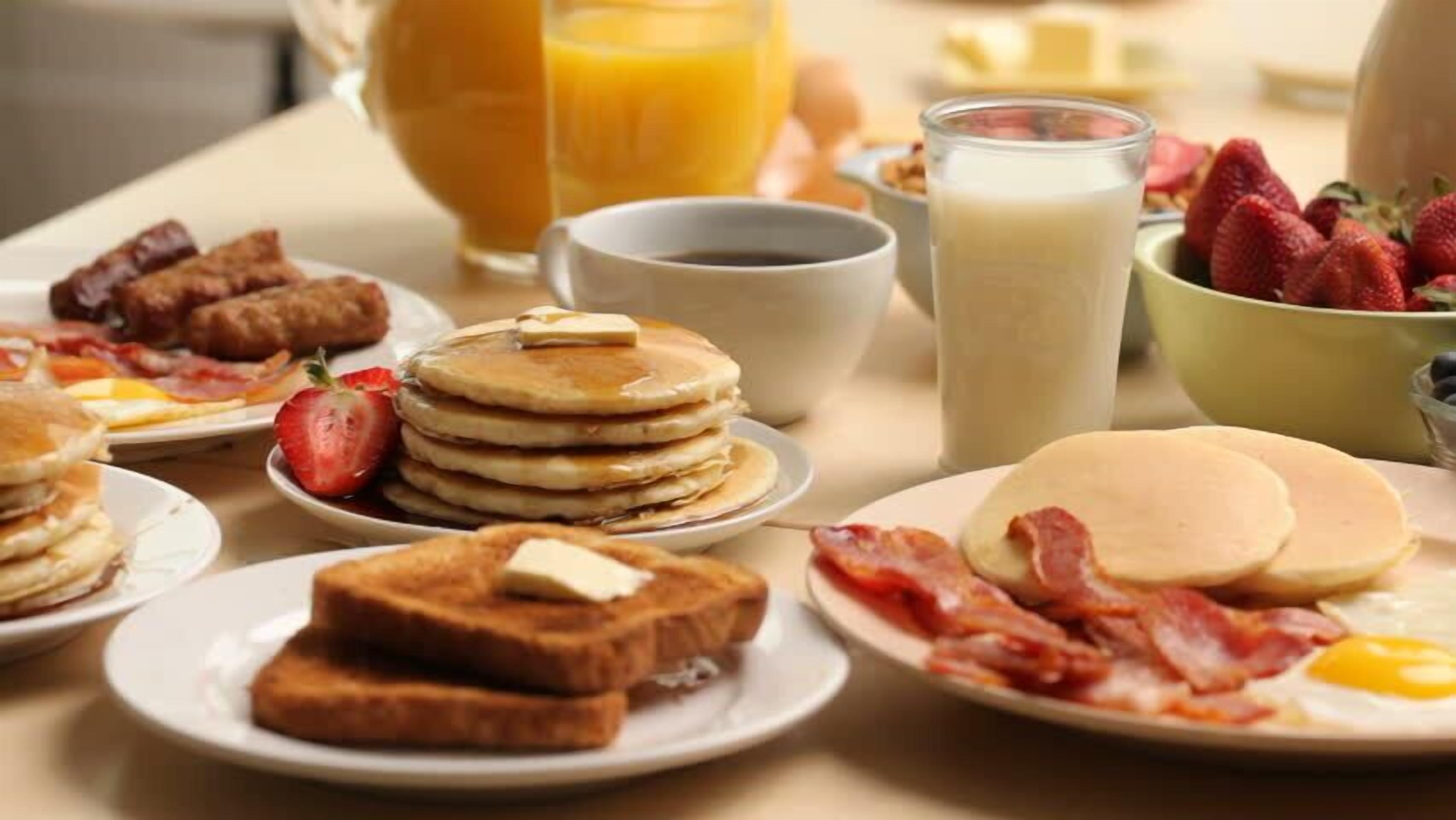 Una mesa de desayuno con panqueques, tocino, huevos, tostadas, fresas y jugo de naranja.
