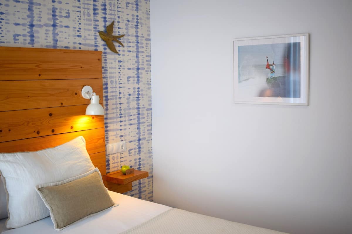 A bedroom with a wooden headboard, a white pillow, a beige pillow, a small wooden nightstand with a green apple, and a framed picture on the wall.