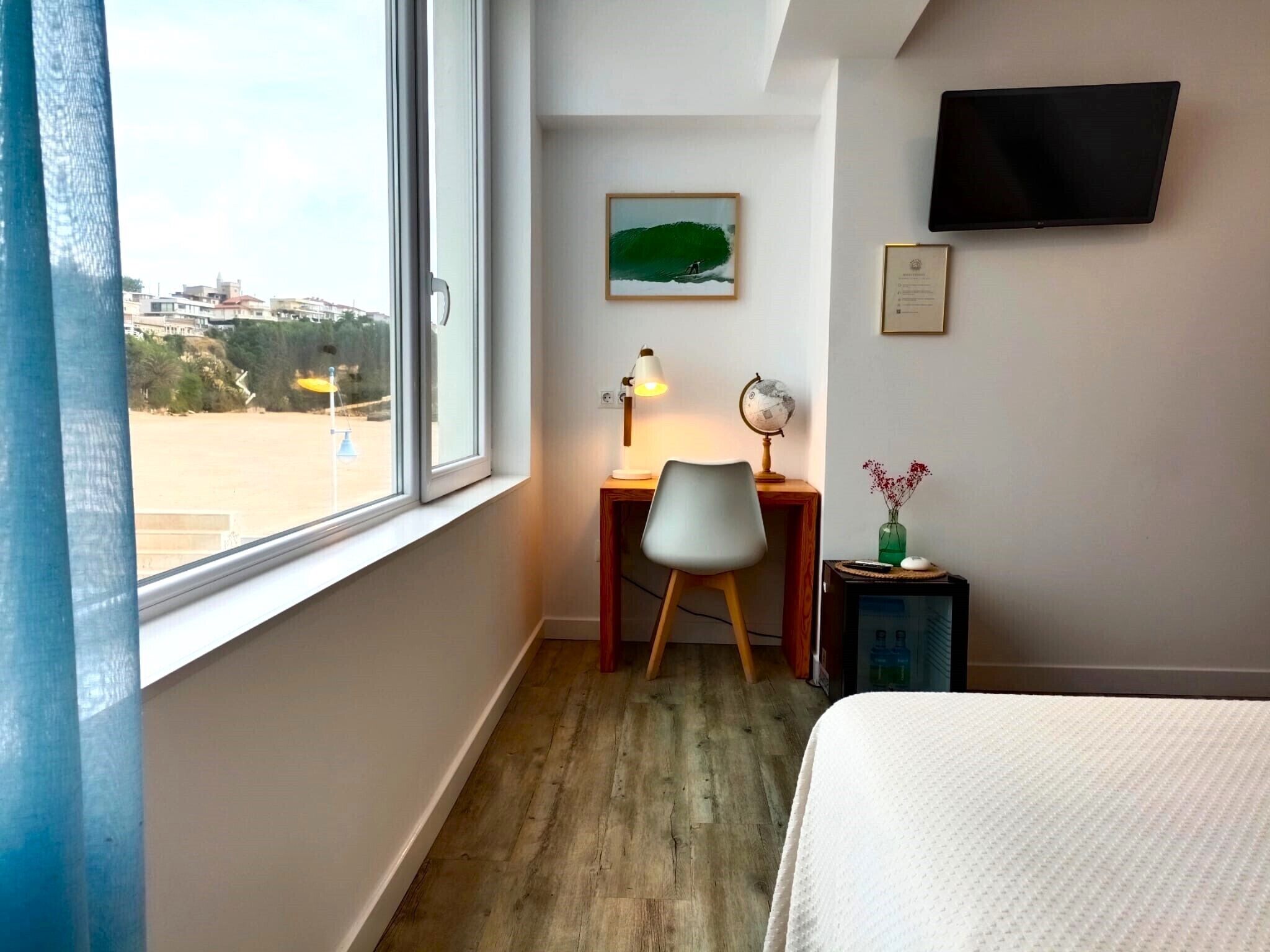 Hotelkamer met een groot raam, een bureau met stoel, een tafeltje met een wereldbol en lamp, een televisie aan de muur en een bed met witte lakens.