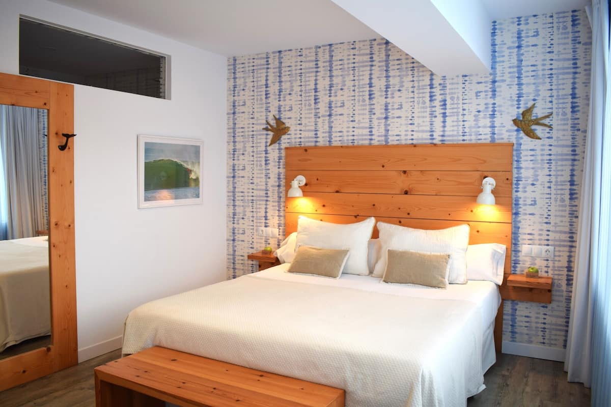 A bedroom with a wooden headboard, a bed with white bedding, a bedside table with a lamp, and a small shelf with a plant.