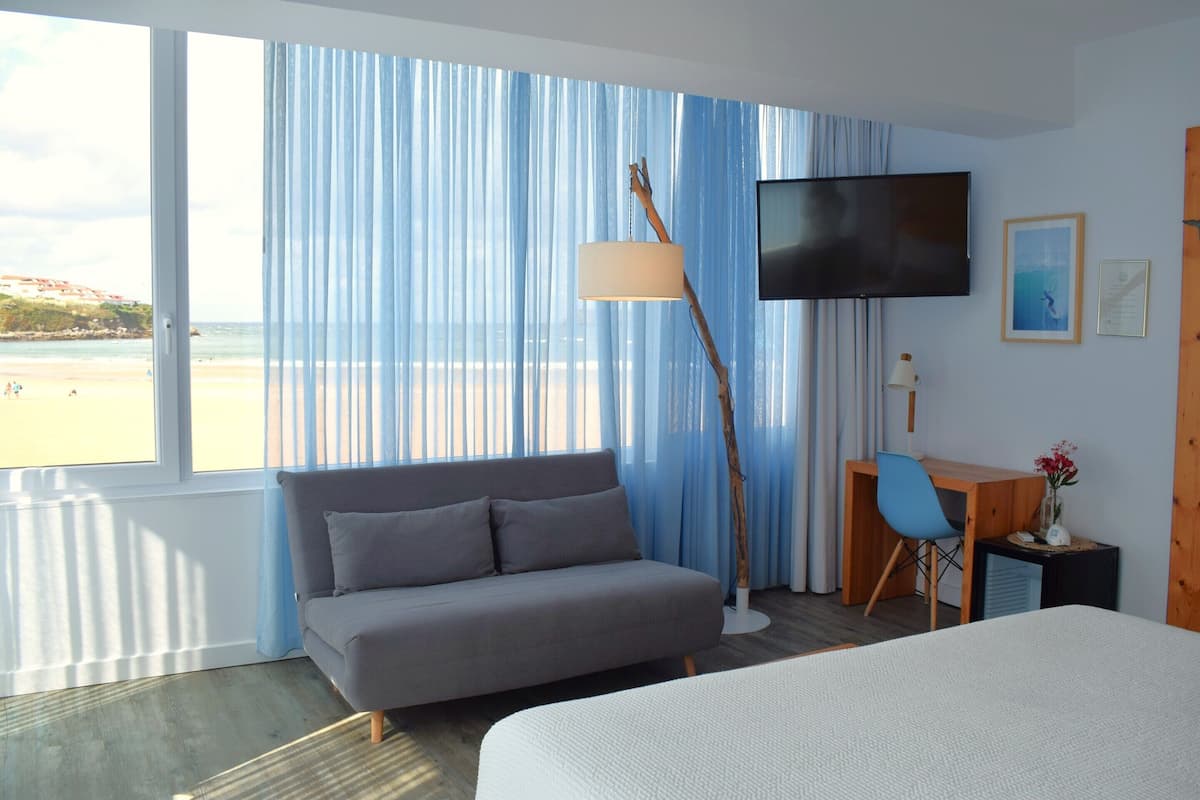 A hotel room with a bed, a sofa, a desk, a TV, and a view of the beach.