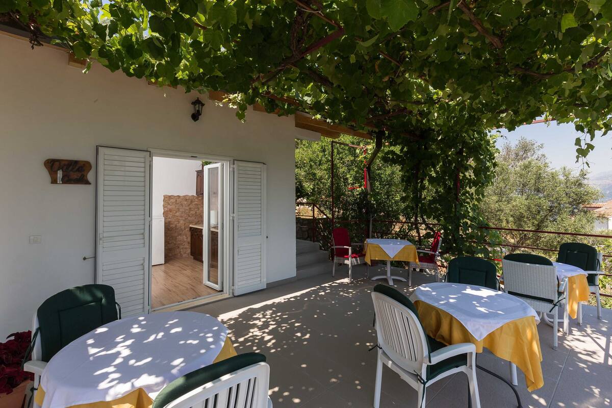 Un espace extérieur aménagé en terrasse, avec des tables et des chaises, une porte-fenêtre coulissante donnant sur l’intérieur et une vue sur le paysage environnant.