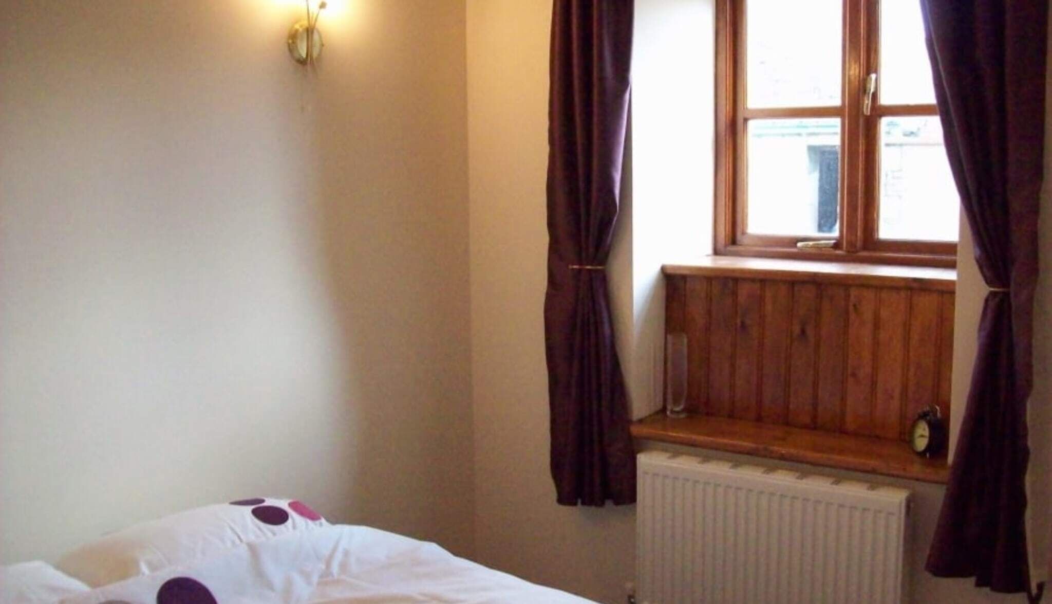 A bedroom with a single bed, a window with curtains, a radiator, and a wall-mounted light fixture.