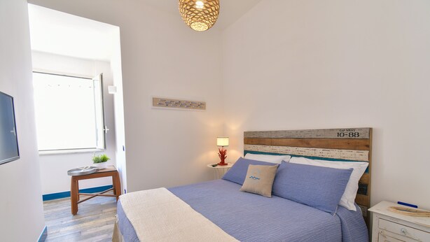 A bedroom with a bed, a nightstand, a lamp, a small table with a plant, and a window. - Rosanna Maison B&B (Ischia)