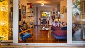 Una sala de estar moderna con sofá, sillón y mesa de centro. Hay un comedor con sillas y una mesa. La habitación tiene un ventanal con persianas, una puerta con felpudo y un espejo decorativo.