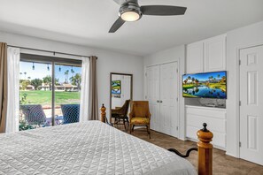 A bedroom with a large bed, a ceiling fan, a television on the wall, and a view of a garden through the window.