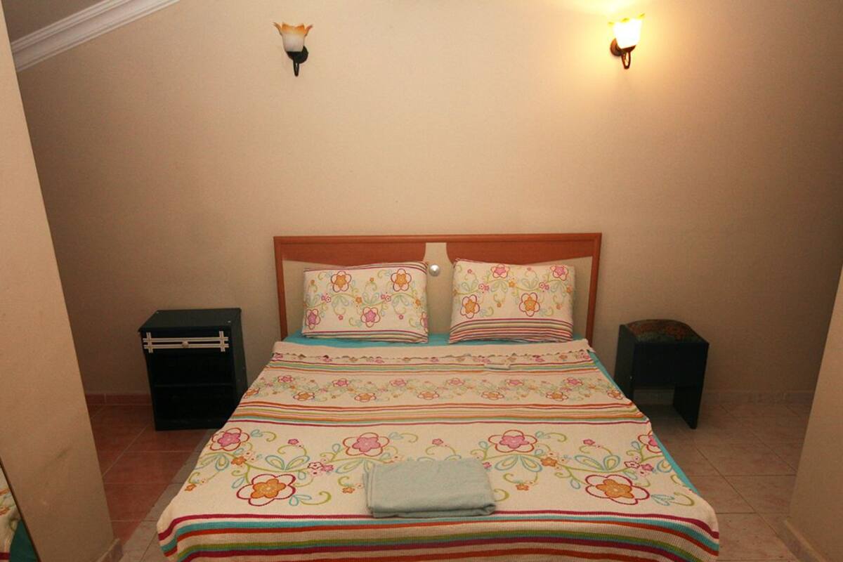 A bedroom with a bed, pillows, a nightstand, and a wall-mounted light fixture.
