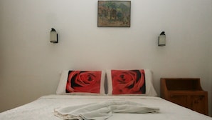 A neatly made bed with a white bedspread and two pillows with red rose designs. There is a small wooden nightstand to the right and a wall-mounted painting above the bed.