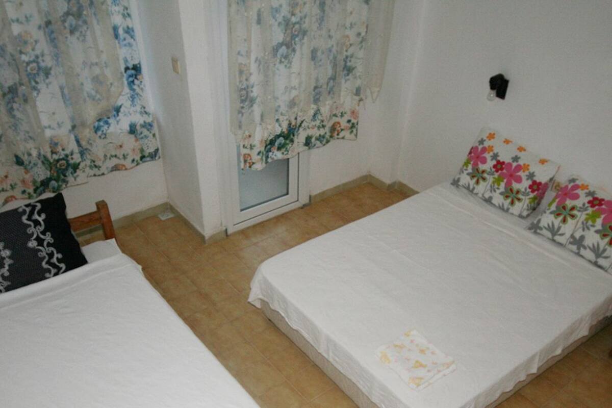 A room with two single beds, a window with floral curtains, a door, and a wall-mounted light fixture.
