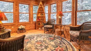 A cozy cabin interior with wooden walls, large windows, a ceiling fan, a fireplace, and wicker furniture. - Walk To The Lake, Beach, Indoor Waterpark And Downtown (Mackinaw City)