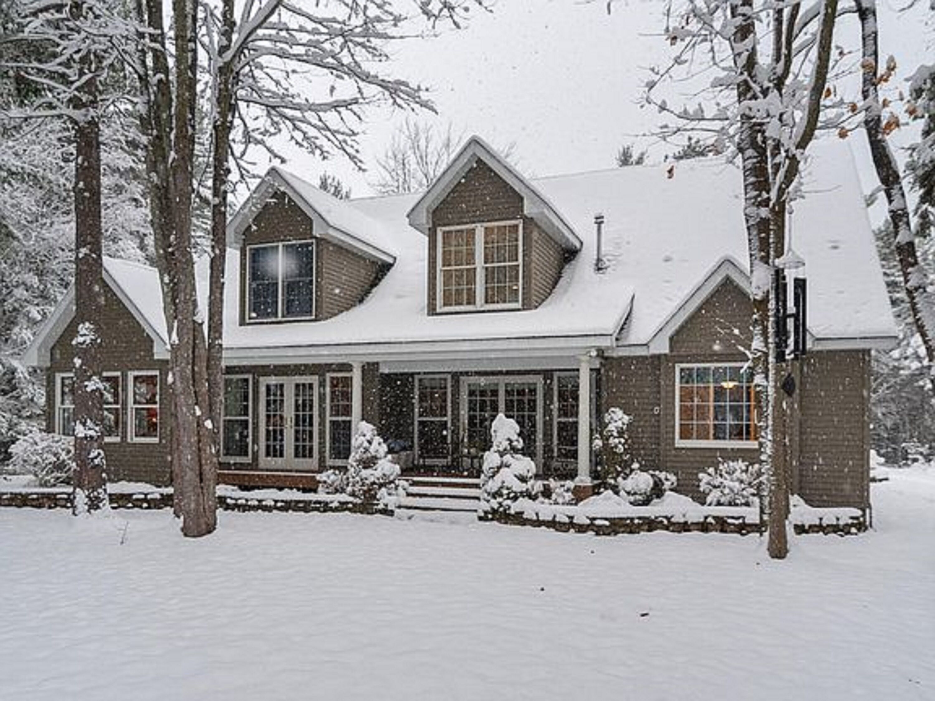 Une maison recouverte de neige, dotée d’une grande véranda à l’avant, de nombreuses fenêtres et d’une façade en briques.