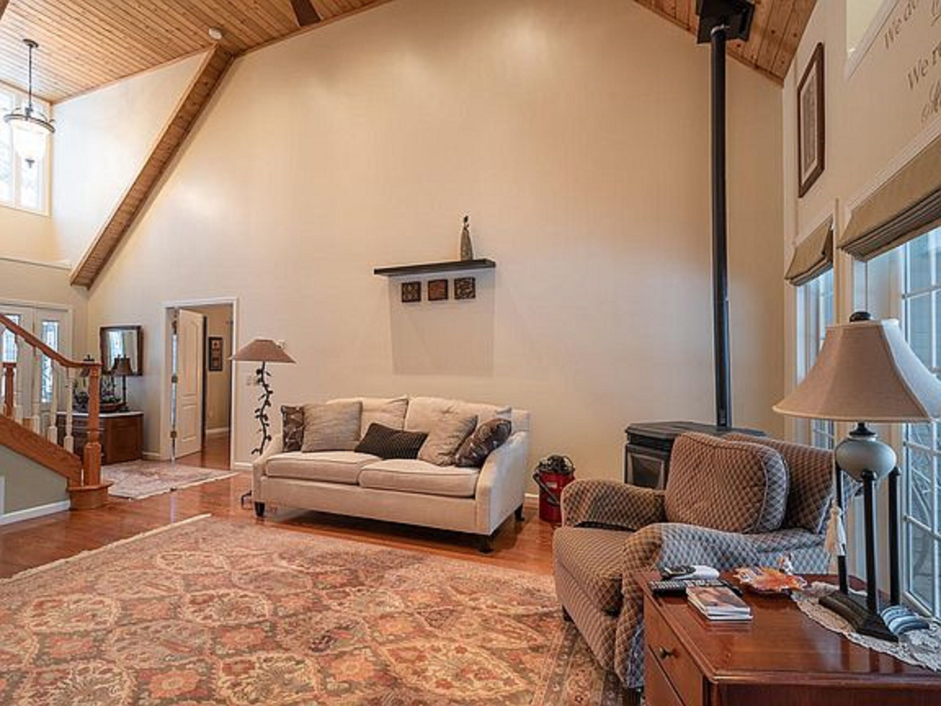 A spacious living room with a high ceiling, a wooden staircase, a beige sofa, a patterned armchair, and a large window providing natural light.