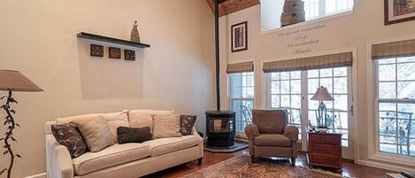 Un salon chaleureux avec un canapé, un fauteuil et un grand tapis. Il y a des fenêtres avec des rideaux, une cheminée et un plafond avec des poutres en bois.