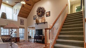 A spacious living room with a wooden staircase, a large window, and a fireplace. - Walk To The Lake, Beach, Indoor Waterpark And Downtown (Mackinaw City)