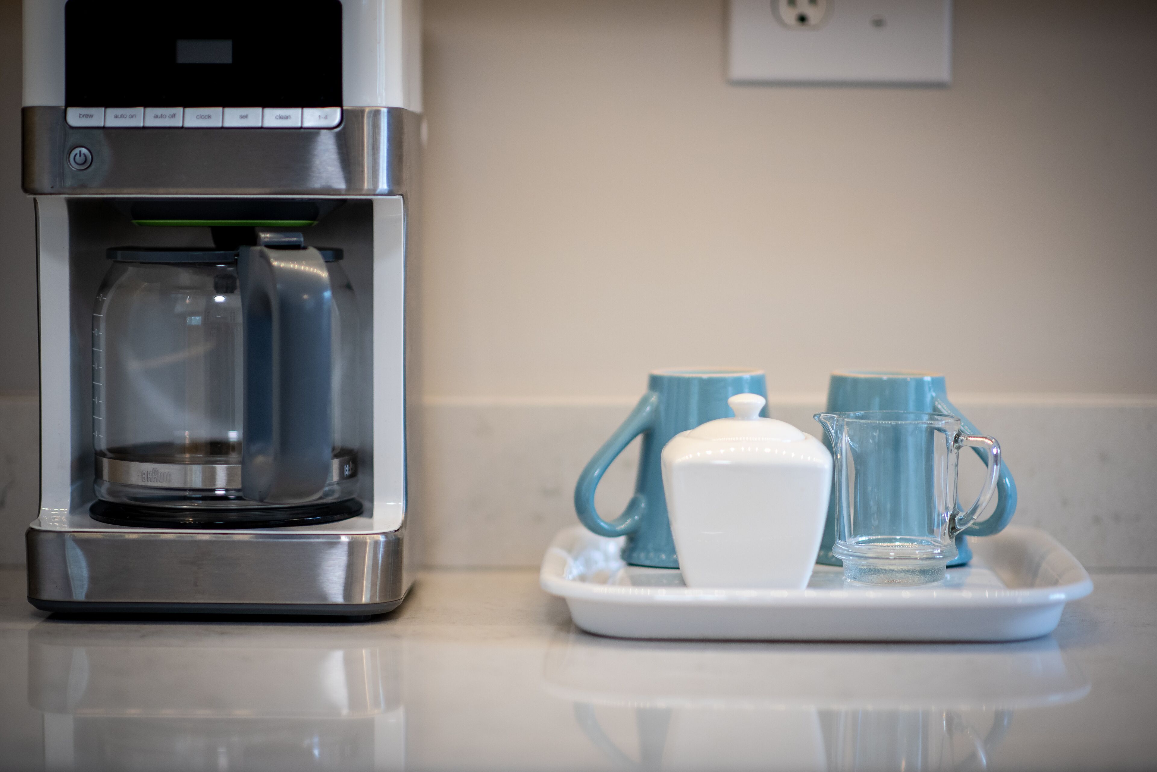 A coffee maker on a countertop with a tray containing a white cup and two blue mugs.