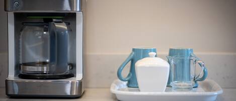 A coffee maker on a countertop with a tray containing a white cup and two blue mugs.