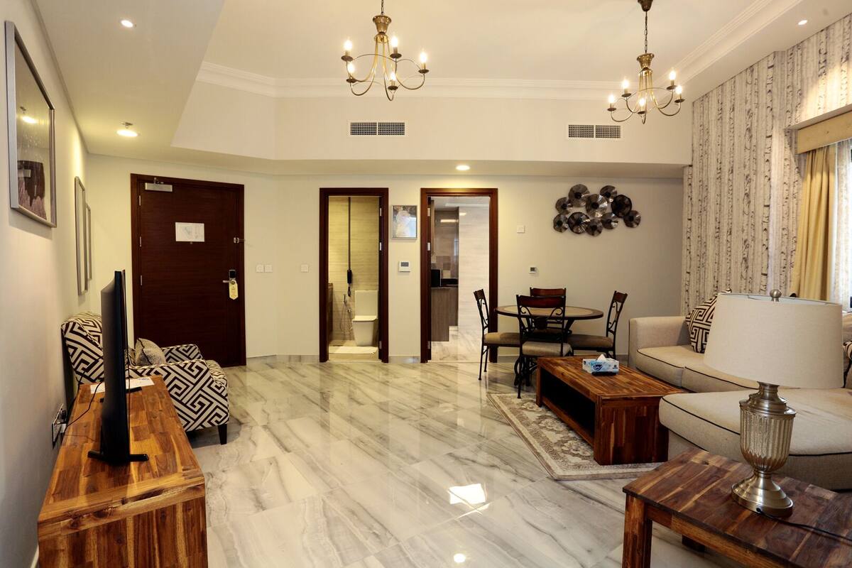 A spacious living room with a marble floor, a sofa, a coffee table, and a TV on a wooden stand. There is a dining area with a table and chairs, and a chandelier hanging from the ceiling.
