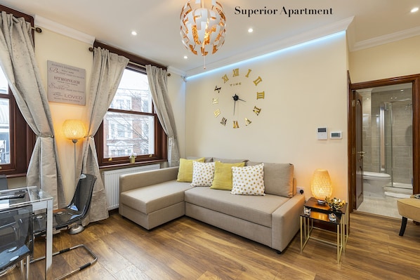A modern living room with a sectional sofa, a glass coffee table, a chair, a wall clock, and a lamp. - Kensington Olympia (London)